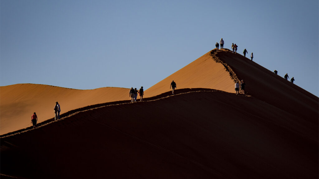 Tegelröda sanddyner där en rad med turister ses gå längs dynernas ryggar.