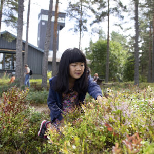 En flicka med svart hår sitter i blåbärsris i skogen utanför naturum.