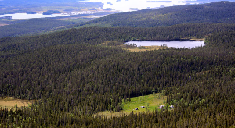 Flygbild med vy över skog och en fäbod i bakgrunden syns en sjö.