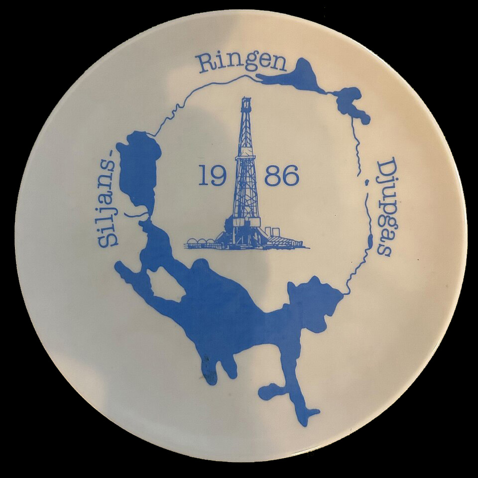 En vit minnestallrik i keramik med en blå siluett av Siljansringens sjöar och en oljerigg i mitten. "Siljansringen Djupgas 1986" står i text på tallriken.