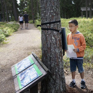 En pojke med svart hår och glasögon läser en fråga till skattjakten utanför naturum.